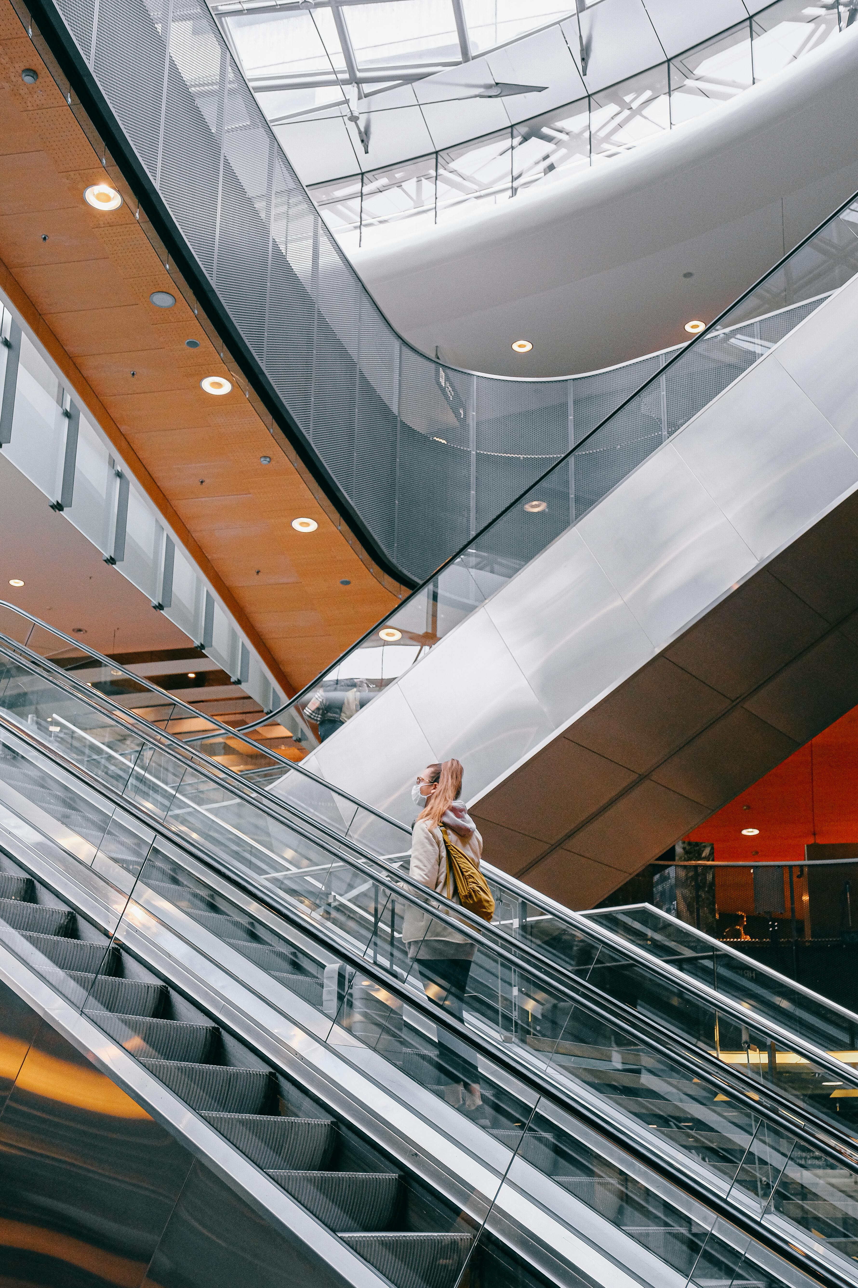 A woman wearing a face mask rides an escalator in a modern airport terminal, emphasizing travel safety.